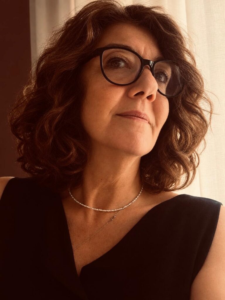 Portrait of a woman with curly brown hair and black-framed glasses, looking to the side in a warm light, wearing a sleeveless black top and a delicate necklace.