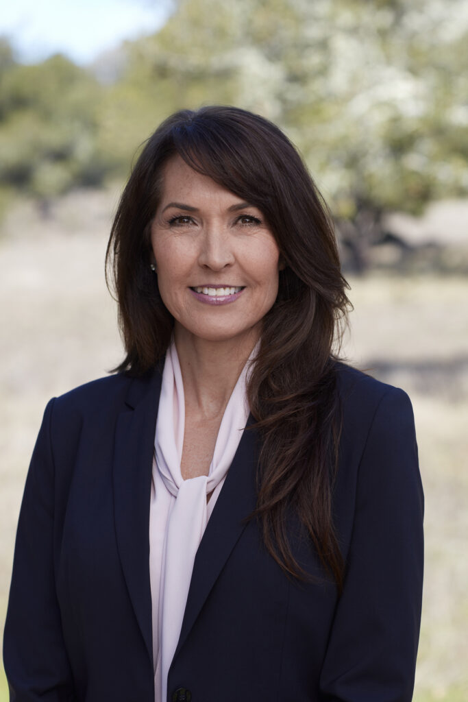 Professional woman in a navy blazer, pale blouse, smiling at the camera outdoors in daylight.