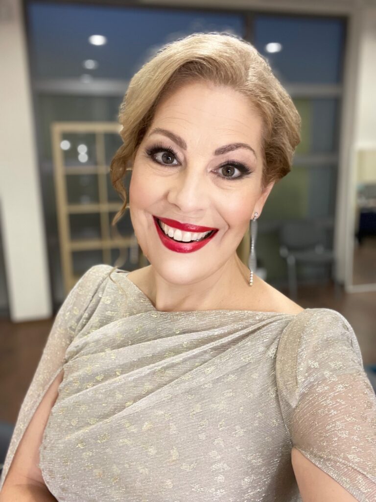 Smiling woman with short blonde hair in a glittery gold dress, wearing red lipstick, posing for a selfie indoors.